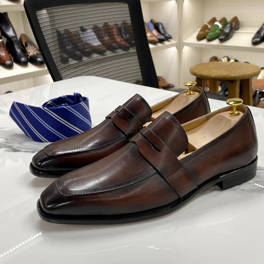 Brown leather loafers with gold accents on a white surface, with a background of more shoes.